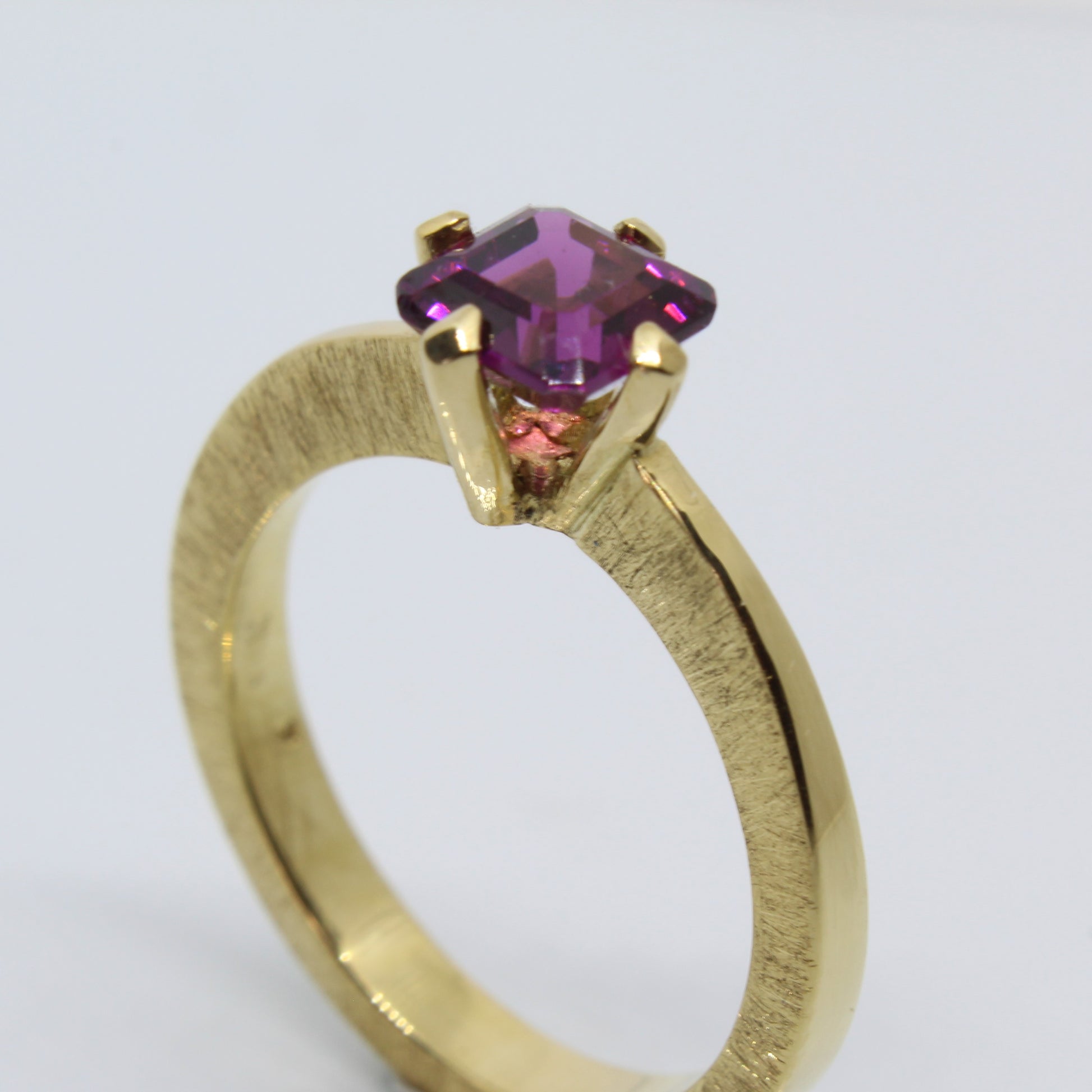 A handmade 18ct yellow gold ring featuring an emerald-cut rhodolite garnet in a secure claw setting. The band showcases a distinctive scored pattern that enhances the elegant lines leading to the garnet.