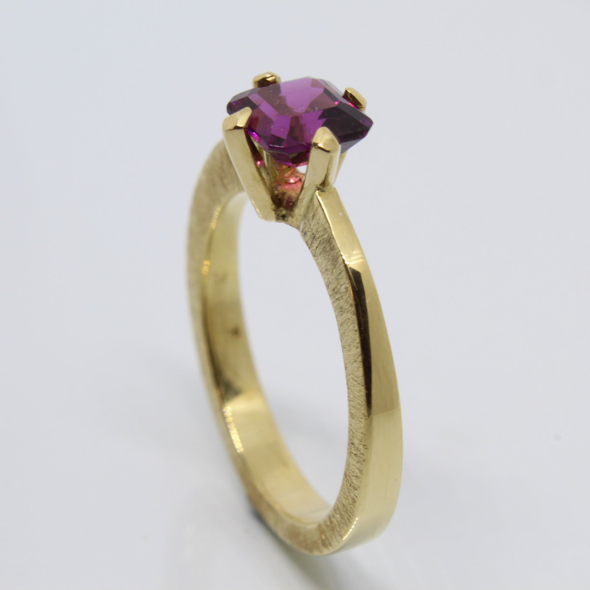 A handmade 18ct yellow gold ring featuring an emerald-cut rhodolite garnet in a secure claw setting. The band showcases a distinctive scored pattern that enhances the elegant lines leading to the garnet.