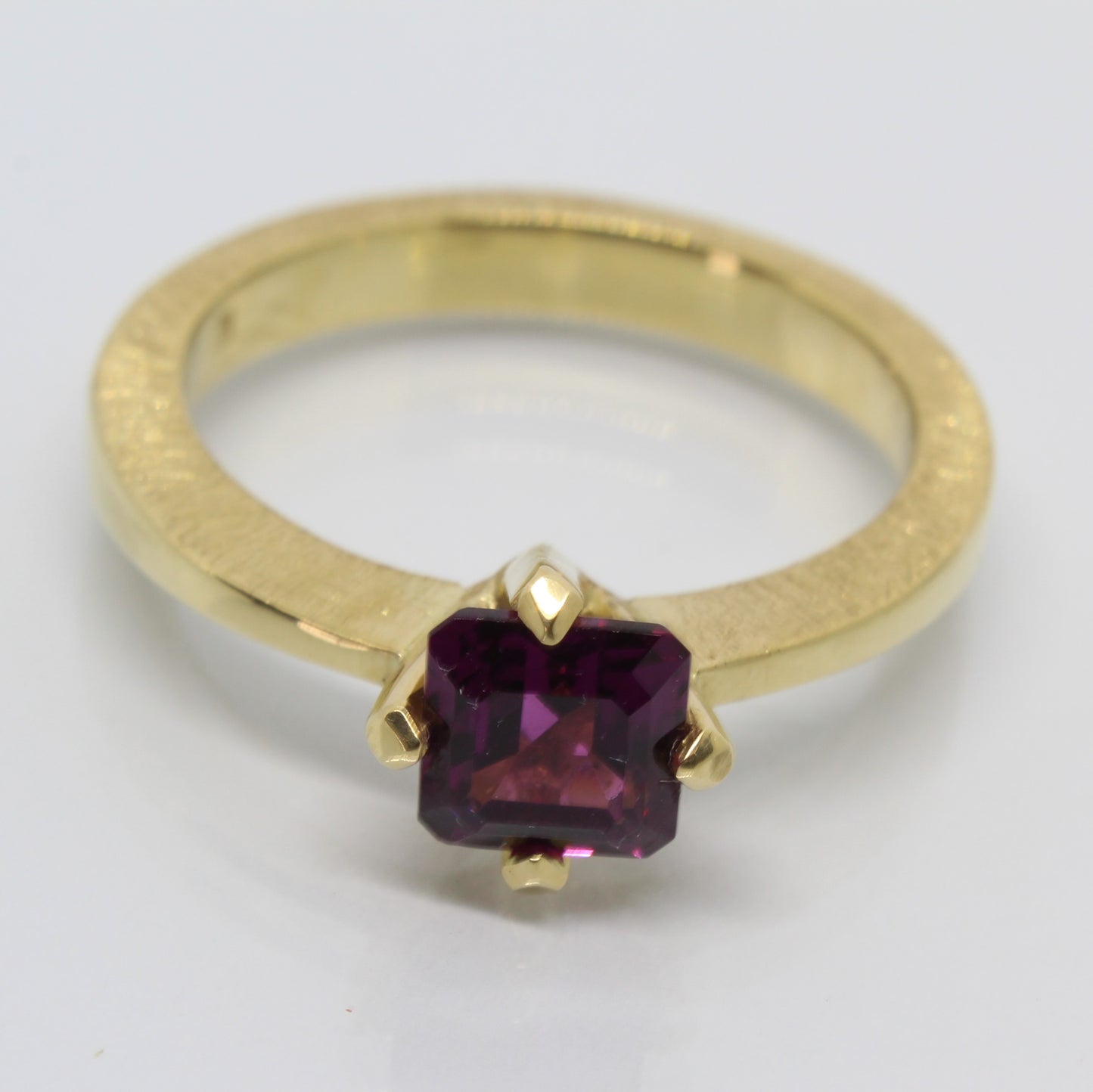 A handmade 18ct yellow gold ring featuring an emerald-cut rhodolite garnet in a secure claw setting. The band showcases a distinctive scored pattern that enhances the elegant lines leading to the garnet.