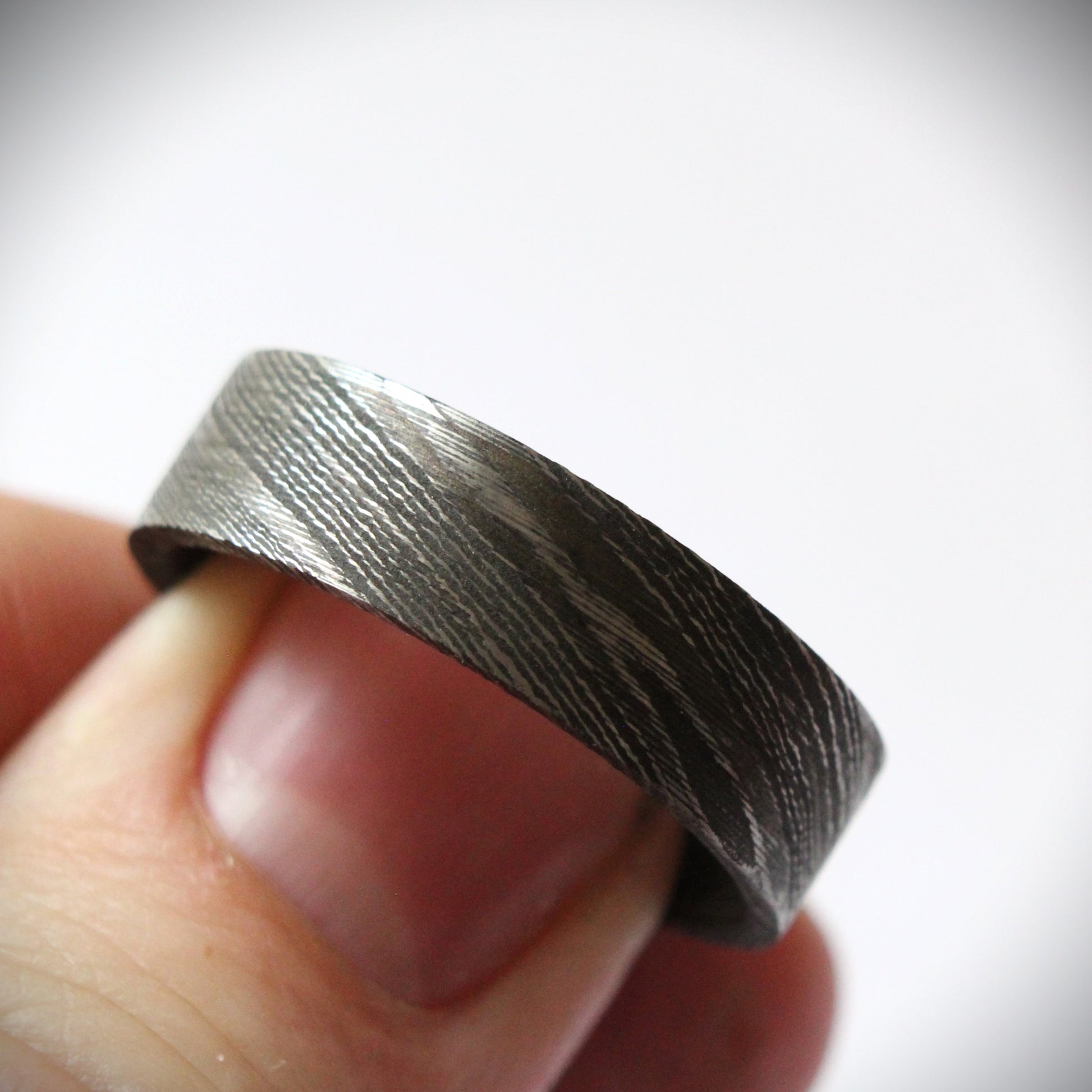 Stunning Damascus steel ring with intricate flowing patterns, heat-treated and blackened for a bold, modern look. The outer band features a unique, layered design with a dark, matte finish, combining strength and elegance in a one-of-a-kind piece of fine craftsmanship. Perfect for a wedding band, engagement ring, or statement piece.