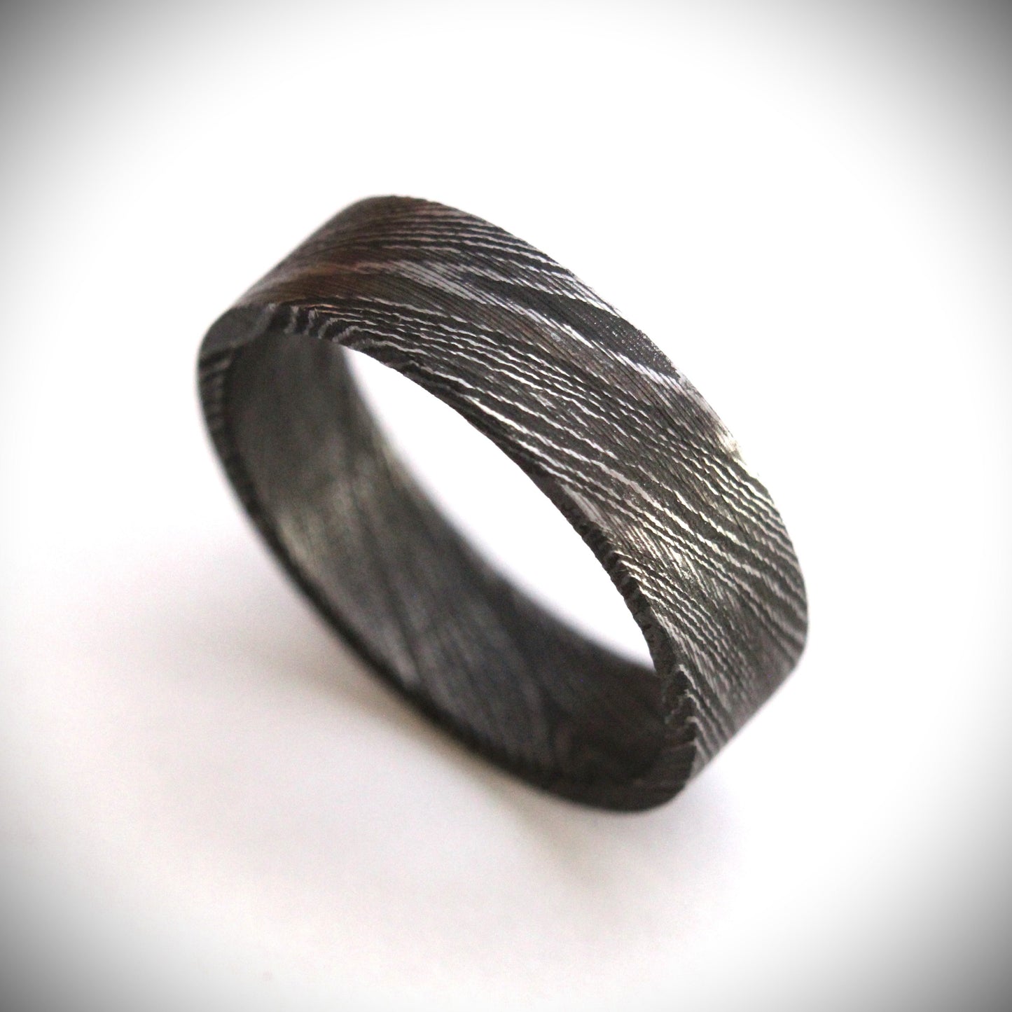 Stunning Damascus steel ring with intricate flowing patterns, heat-treated and blackened for a bold, modern look. The outer band features a unique, layered design with a dark, matte finish, combining strength and elegance in a one-of-a-kind piece of fine craftsmanship. Perfect for a wedding band, engagement ring, or statement piece.